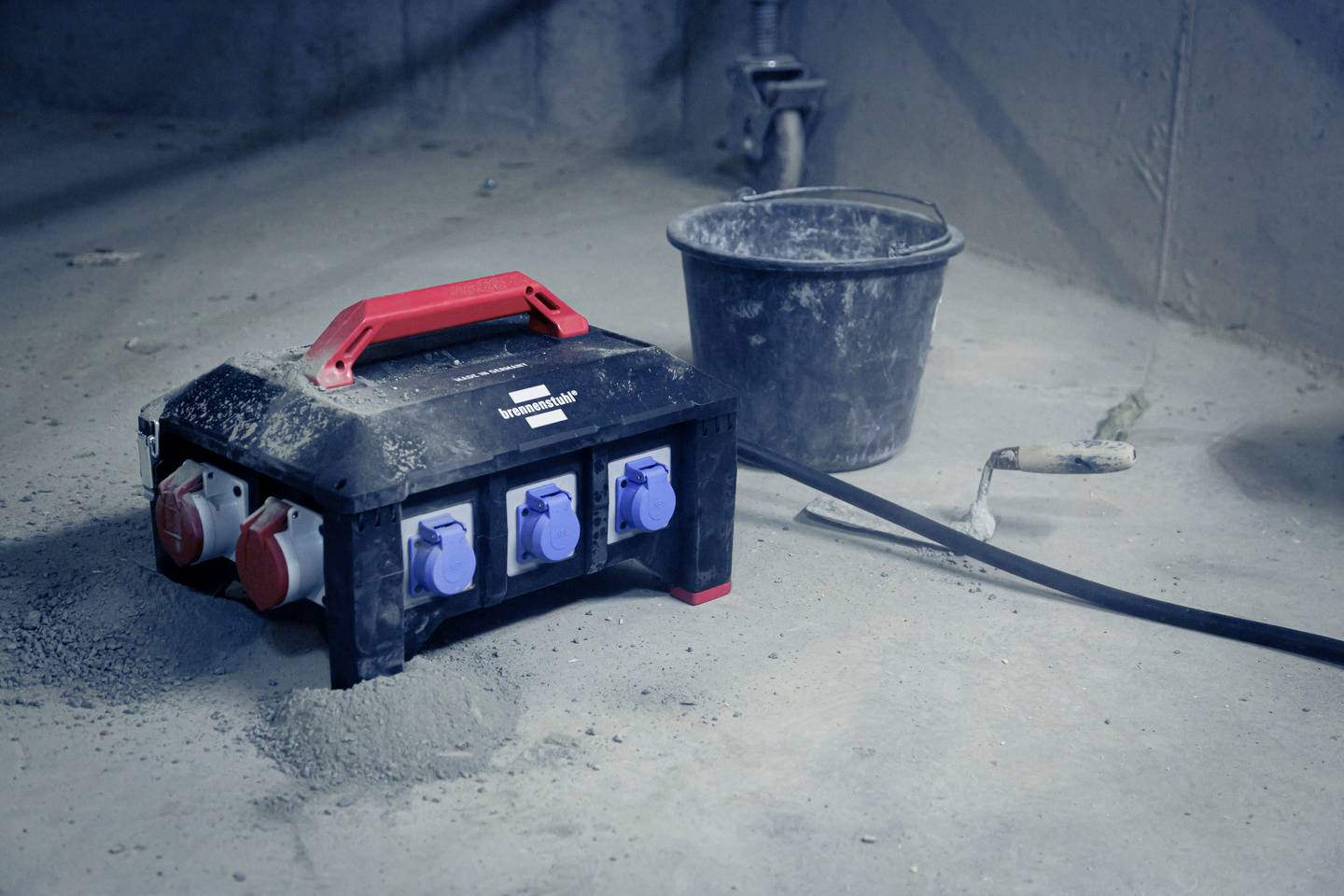 Eine schwarze Kabeltrommel mit mehreren blauen und roten Steckdosen, daneben ein grauer Eimer und ein Werkzeug auf betoniertem Boden.