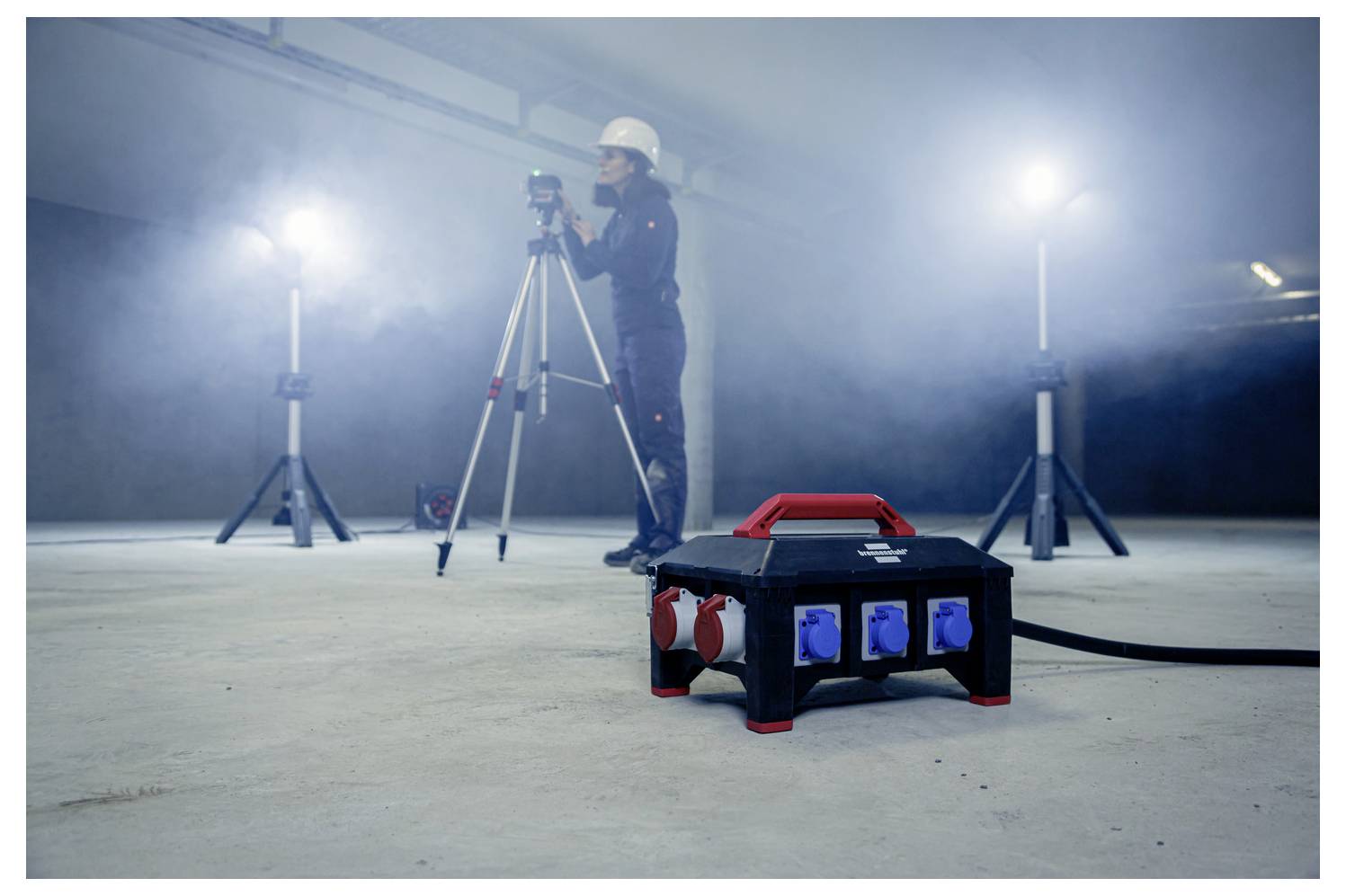 Bauarbeiter misst einen verrauchten Innenraum mit einem auf einem Stativ montierten Laserscanner; tragbare Stromverteilungsbox im Vordergrund.