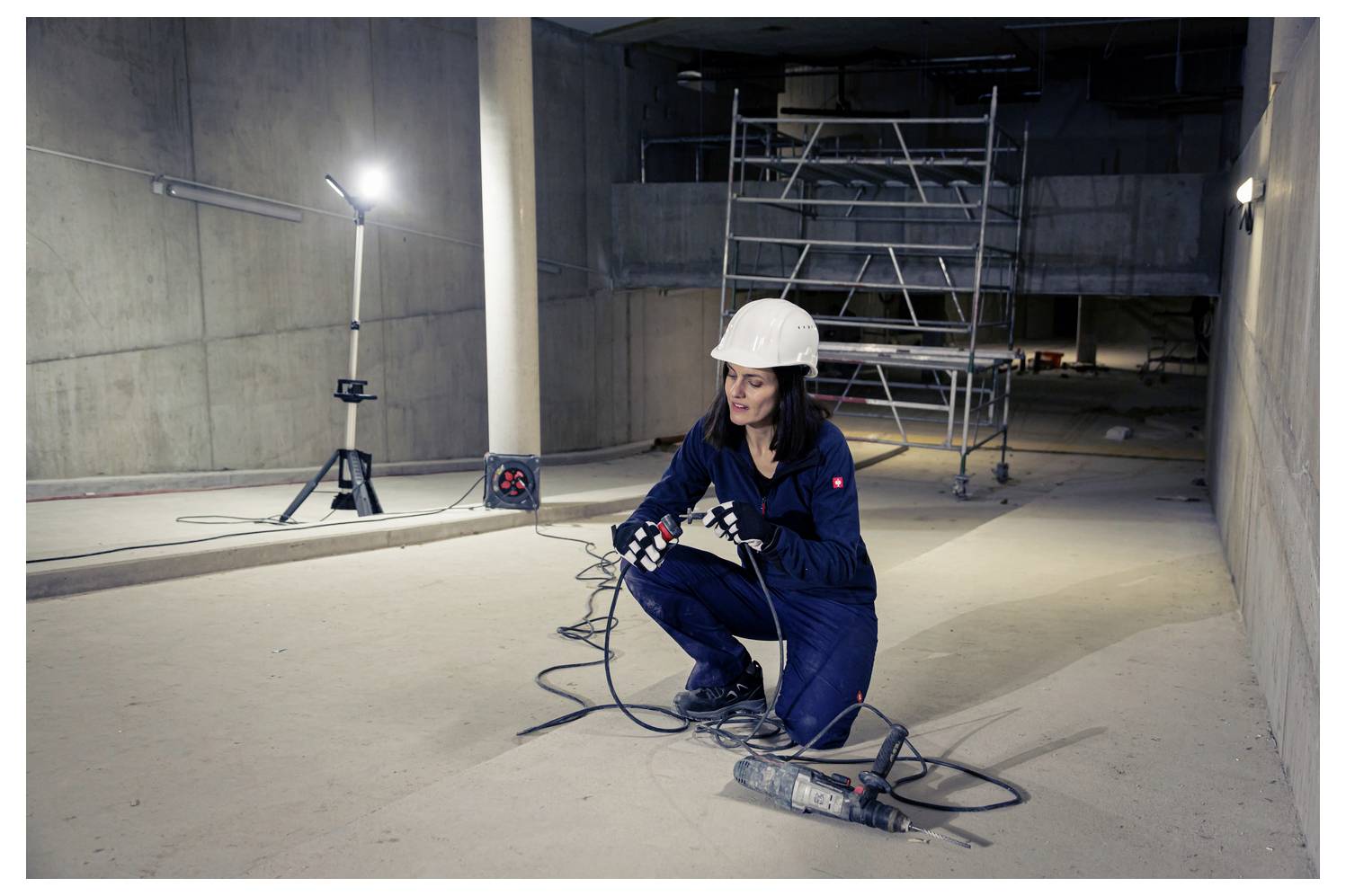 Bauarbeiter mit Schutzhelm hockt auf einem Betonboden und handhabt Stromkabel an einer schwach beleuchteten Baustelle mit Gerüsten hinter ihr.
