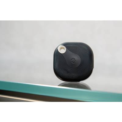 Ein schwarzer, quadratischer Wireless-Tracker mit einem geprägten Logo liegt auf einem Glasregal.