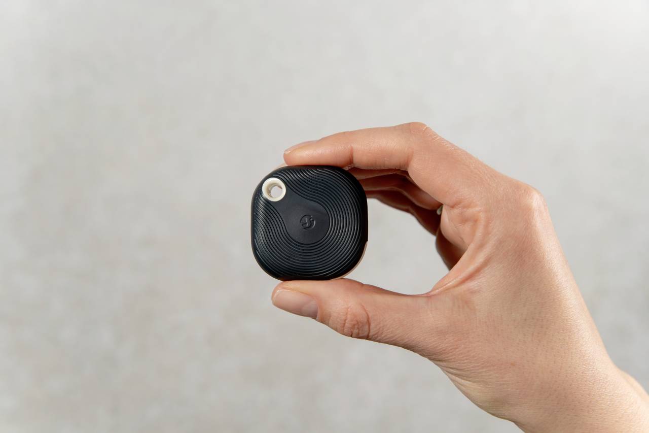 Eine Hand hält einen kleinen, quadratischen, schwarzen Tracker mit einer Linse in der oberen Ecke. Der Hintergrund ist neutral.