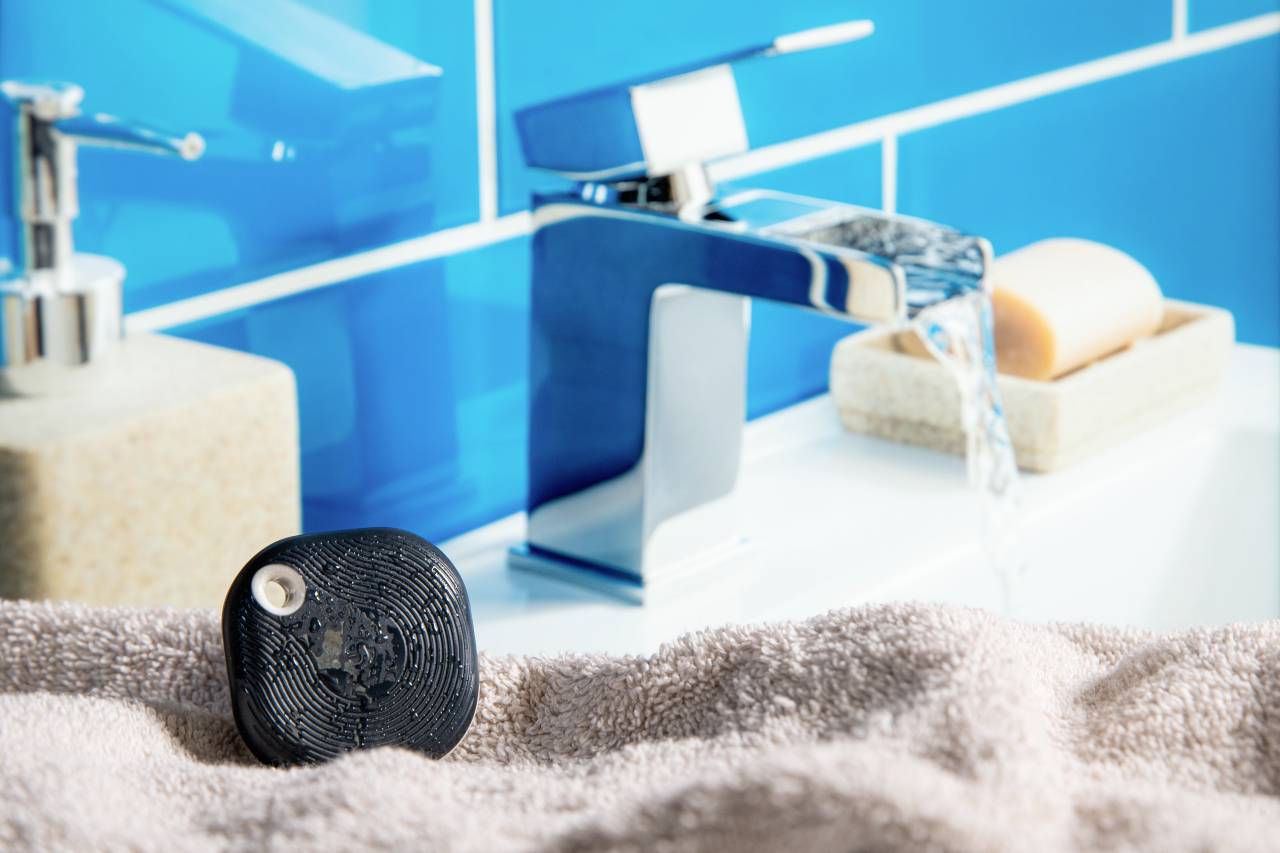 Ein schwarzes Gerät liegt auf einem Handtuch neben einem modernen Wasserhahn im Bad. Im Hintergrund ein Seifenspender und Seife.