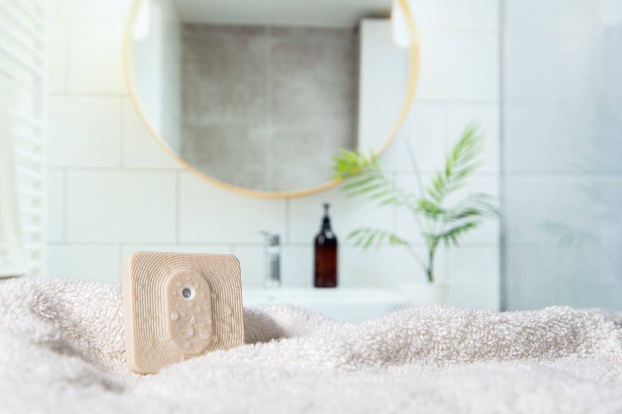 Ein Holzstück mit kleinen Knöpfen liegt auf einem Handtuch. Im Hintergrund ein rundes Spiegel und ein Badezimmer mit Pflanze.