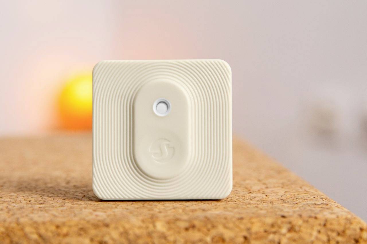 Kleiner quadratischer Sensor mit strukturiertem Gehäuse, zentralem Knopf und Logo, platziert auf einer Korkoberfläche im Vordergrund.