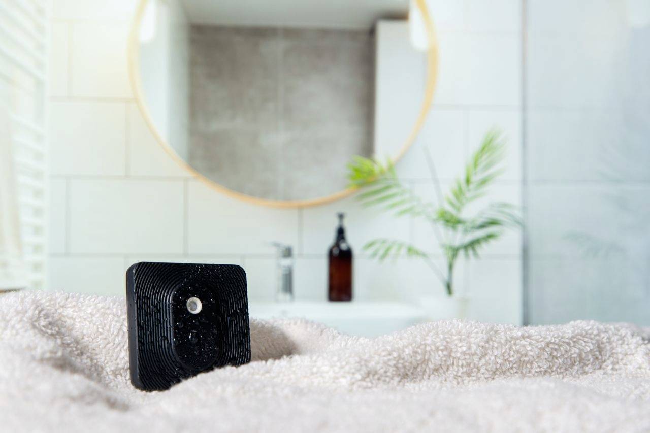 Schwarzer Würfel mit Linse auf Handtuch im Badezimmer. Unscharfer Spiegel und Pflanze im Hintergrund.