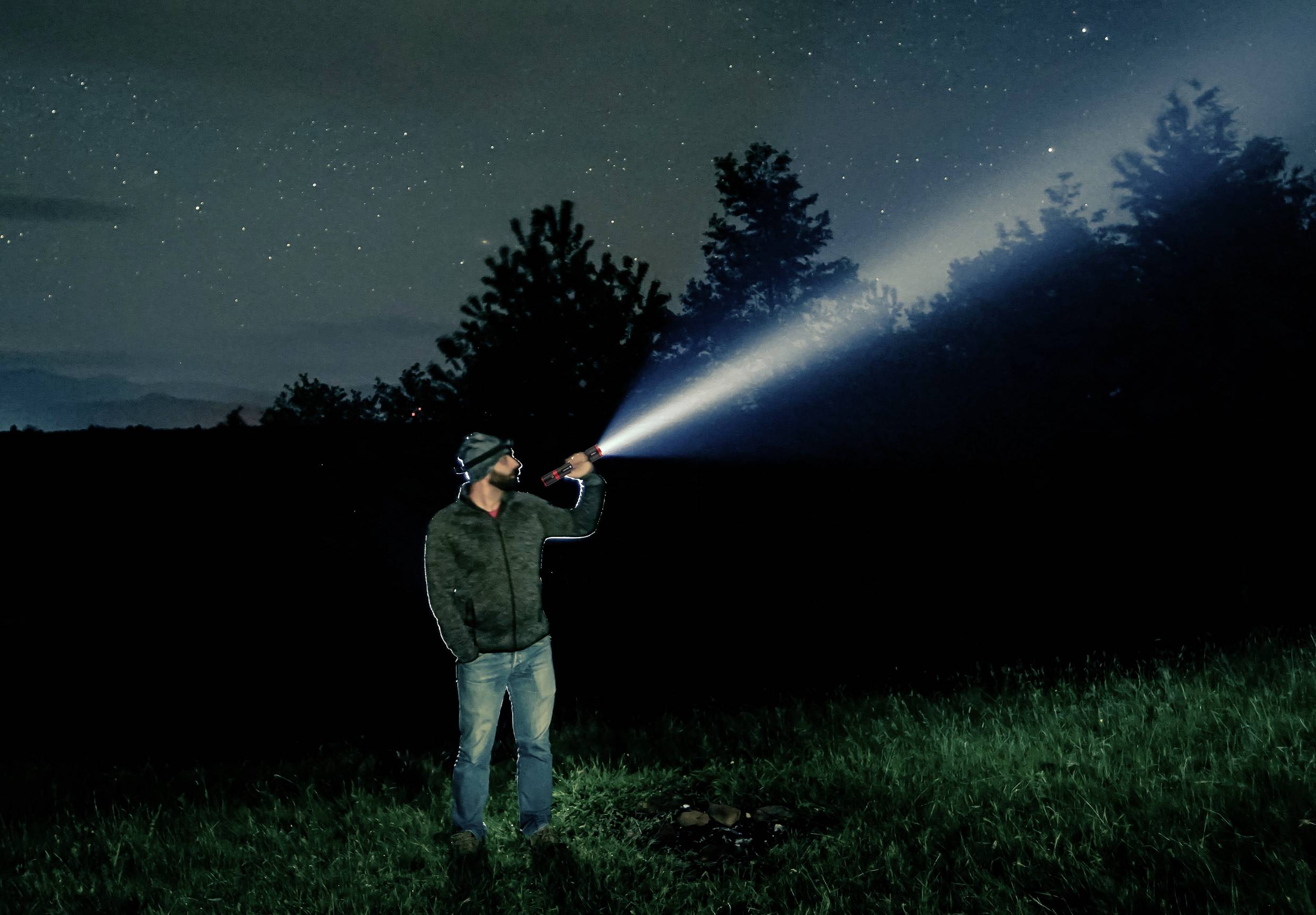 Ein Mann steht nachts im Freien und leuchtet mit einer Taschenlampe in den Himmel. Der Sternenhimmel ist im Hintergrund sichtbar.