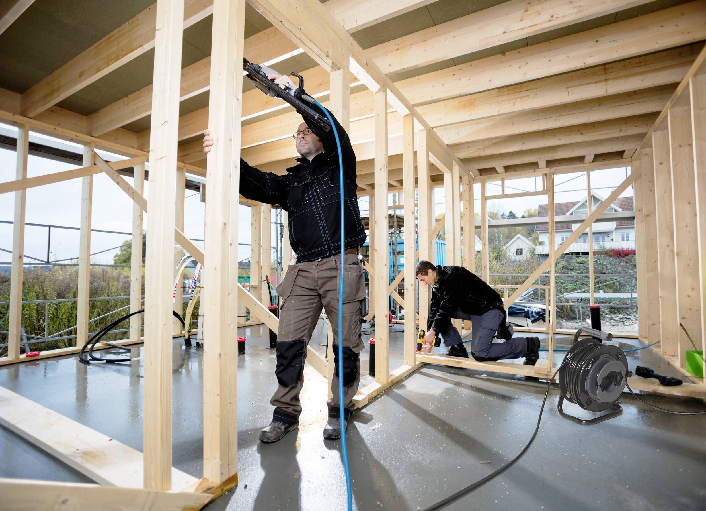 Zwei Personen arbeiten an einem Hausbauprojekt. Eine Person benutzt ein Werkzeug, die andere kniet und arbeitet ebenfalls mit Werkzeug.