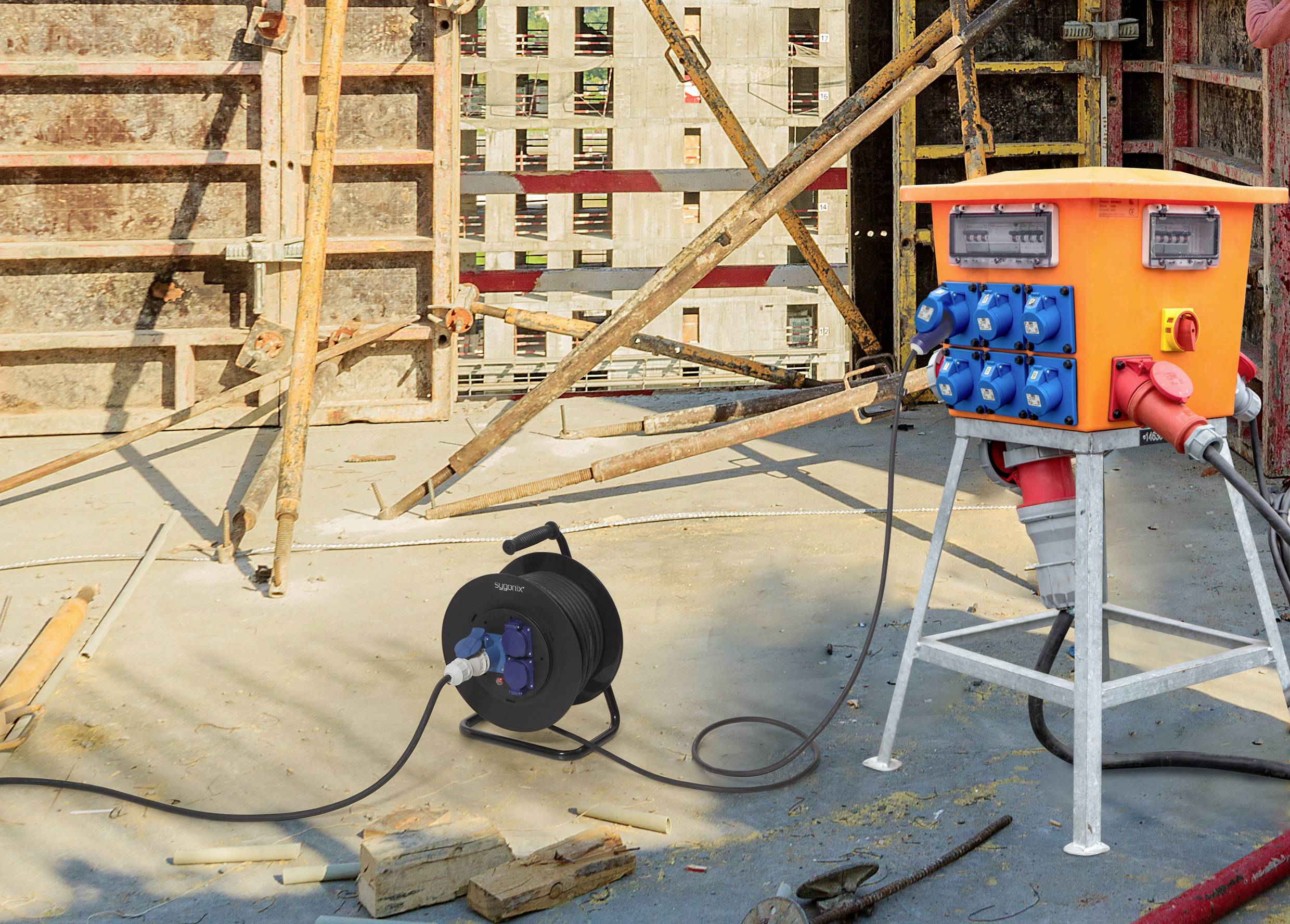 Eine Baustelle mit einem orangefarbenen Stromverteilerkasten und einer Kabeltrommel im Vordergrund, umgeben von Baugerüsten.