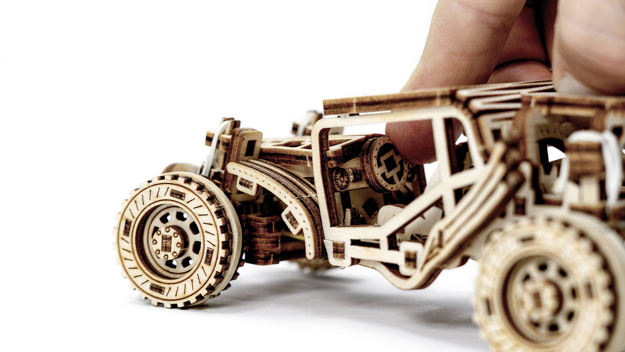 Holzmodell eines Autos mit sichtbaren Zahnrädern und Mechanismen, das von einer Hand berührt wird.