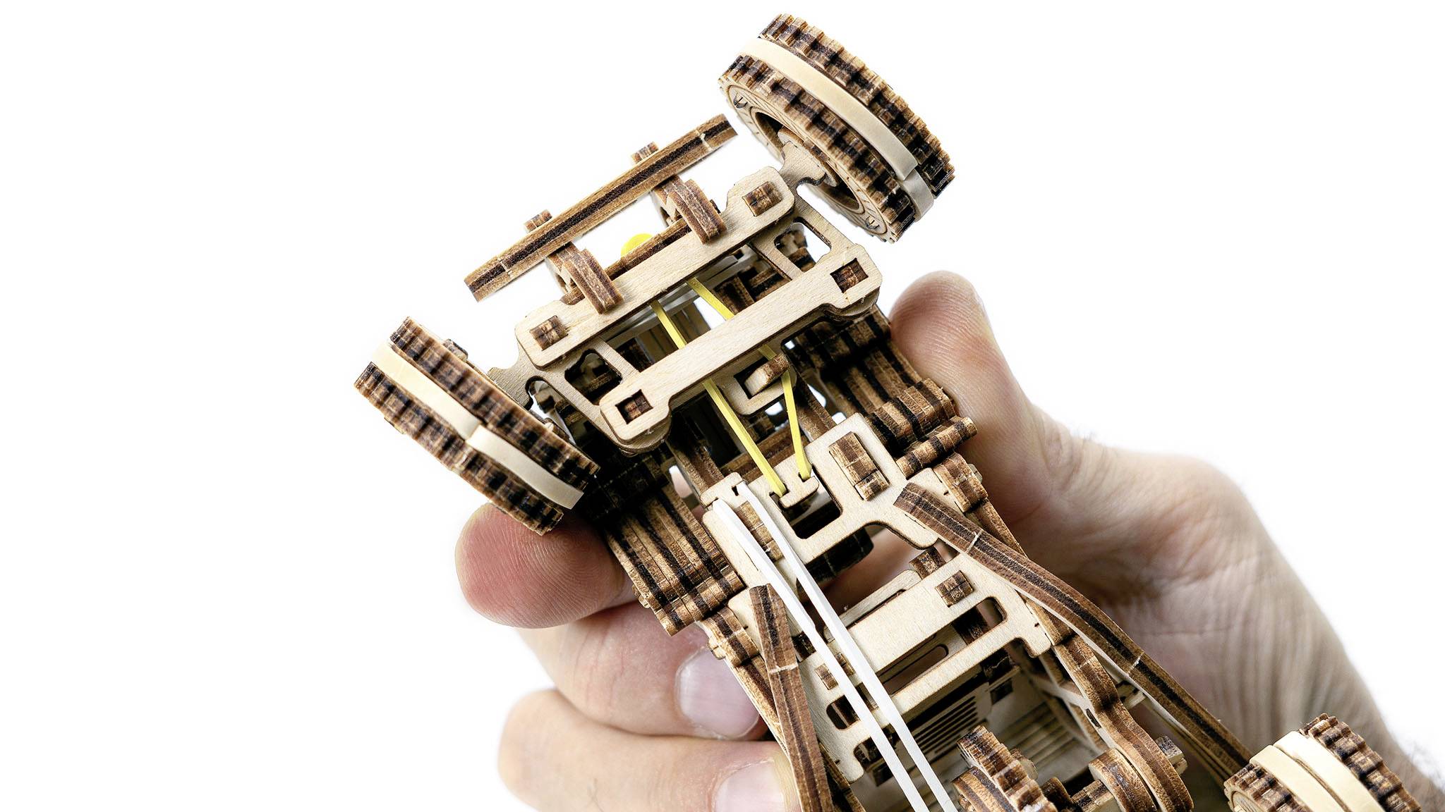 Hand hält ein Miniatur-Rennwagenmodell aus Holz, das die untere Struktur und Zahnräder zeigt. Die Räder sind groß und detailliert.