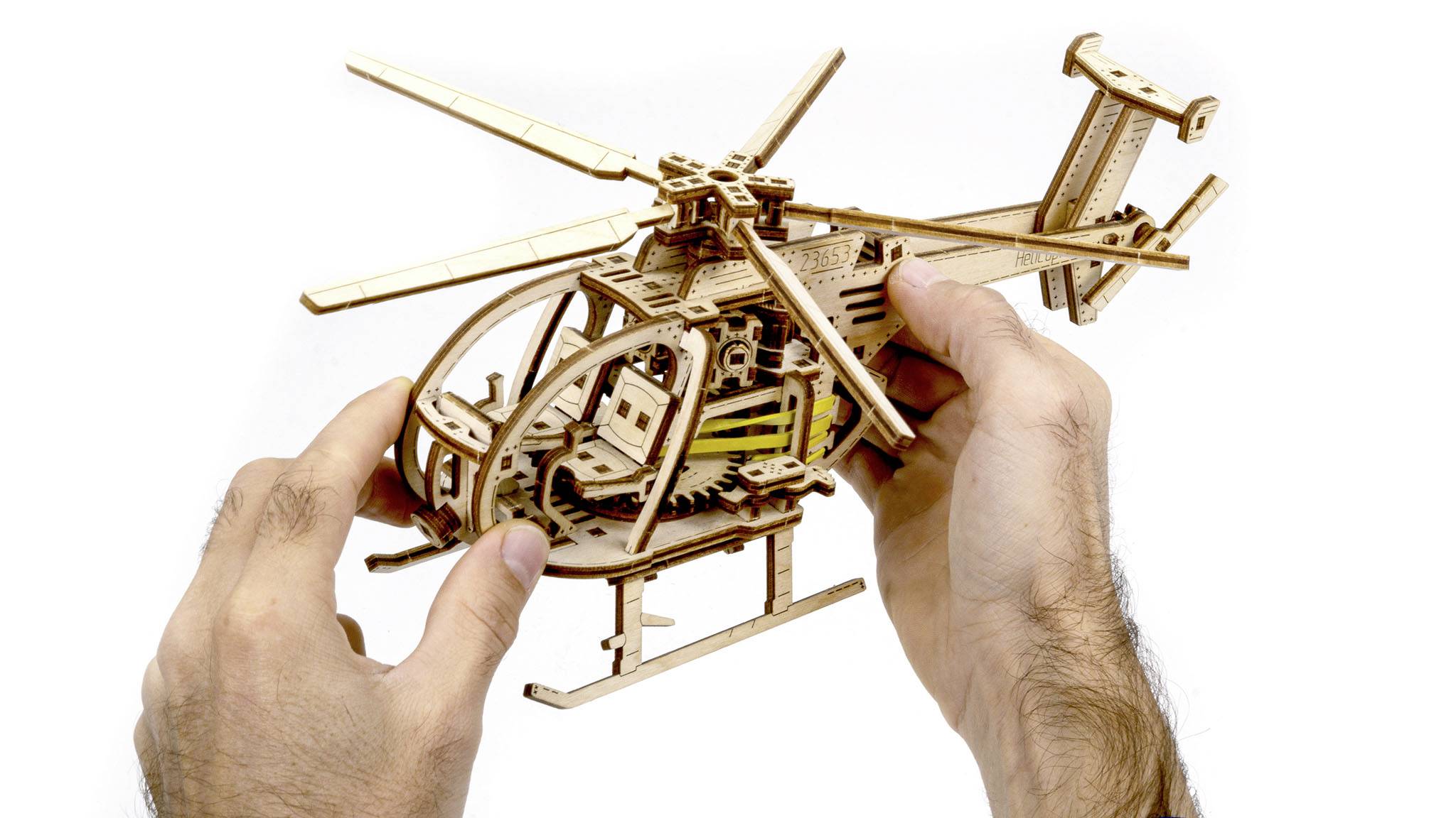 Zwei Hände montieren ein Holzmodell eines Hubschraubers. Das Modell zeigt detaillierte Komponenten und Zahnräder.
