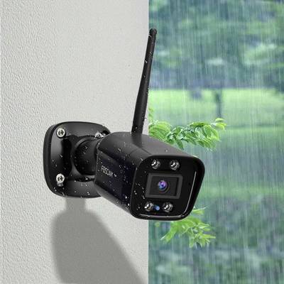 Überwachungskamera an einer Wand im Regen montiert, mit einer Antenne für drahtlose Verbindung. Hintergrund zeigt verschwommene Vegetation.