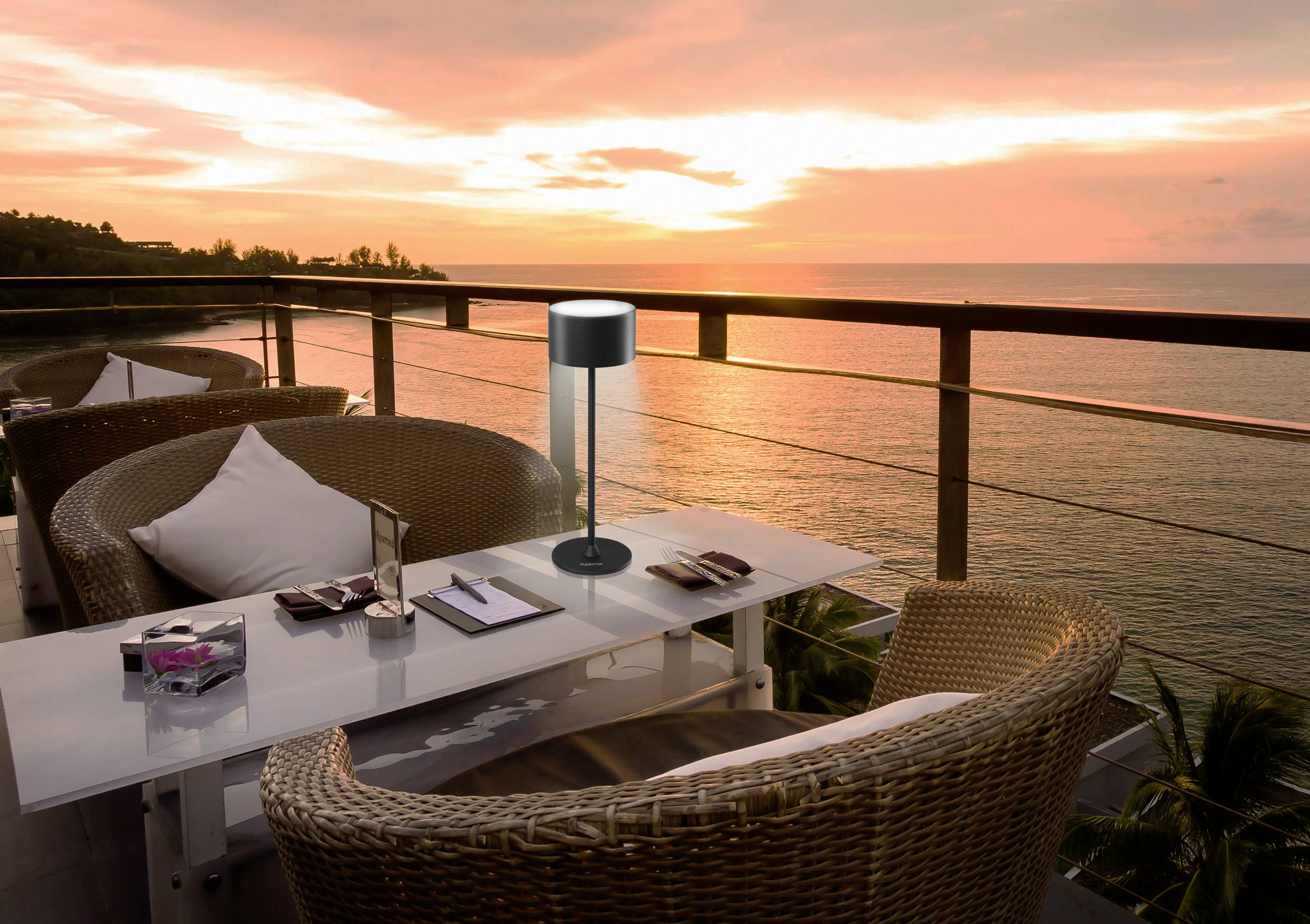 Sonnenuntergang auf einer Terrasse mit Korbstühlen und einem Tisch. Blick auf das Meer mit einer warmen, orangefarbenen Himmelskulisse.