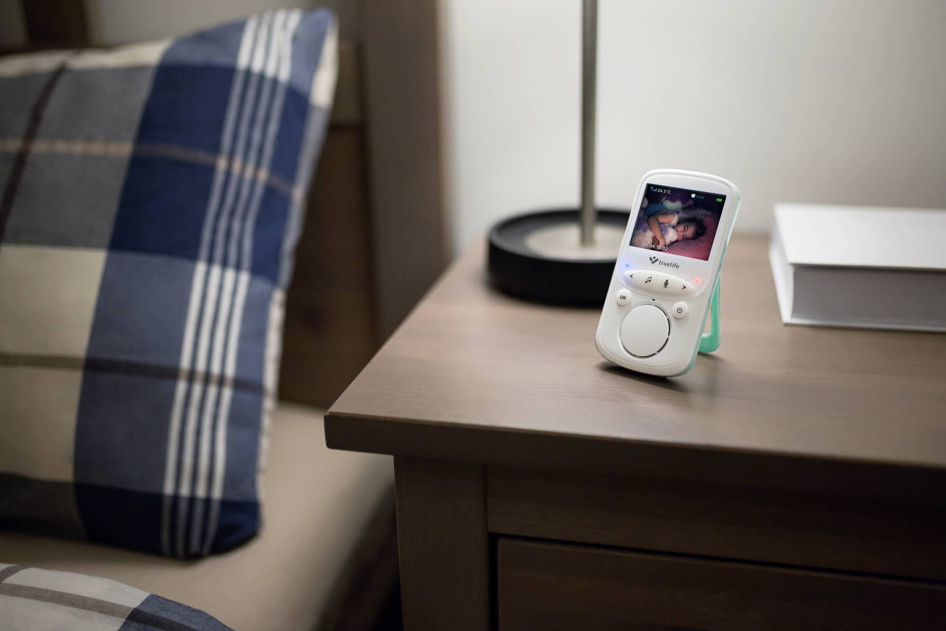 Auf einem Nachttisch steht ein Babyphone mit Bildschirm, das ein schlafendes Baby zeigt. Daneben sind eine Lampe, ein Buch und ein Kissen.