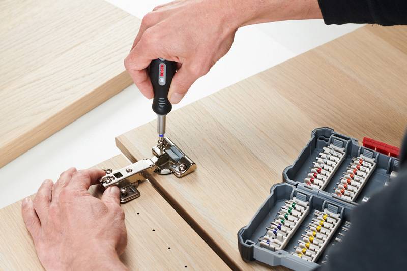 Eine Person montiert mit einem Schraubendreher ein Scharnier an einem Holzmöbelstück. Daneben liegt ein Werkzeugkasten mit Schrauben.