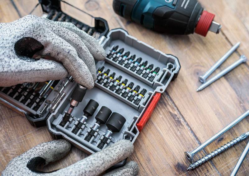 Mechanikerhände in Handschuhen suchen in einem Behälter voller Schraubendreherbits, daneben liegen Schrauben und ein Akkuschrauber.