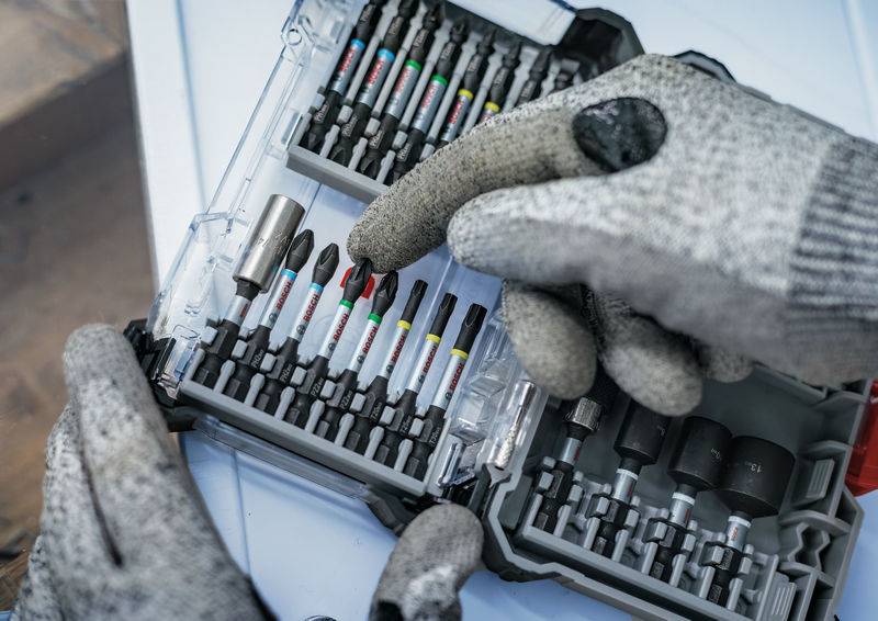 Eine Person mit grauen Handschuhen wählt einen Schraubendreheraufsatz aus einem offenen Set mit verschiedenfarbigen Aufsätzen.