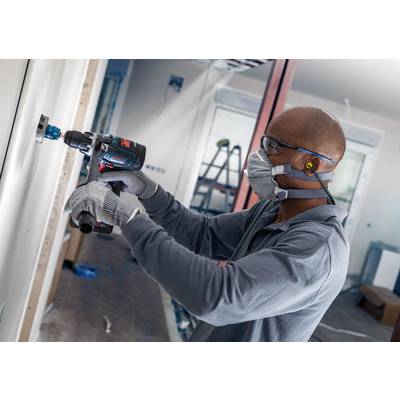 Eine Person bohrt ein Loch in eine Wand in einem Haus, trägt Schutzbrille, Handschuhe und Maske. Renovierungsarbeiten sind im Gange.