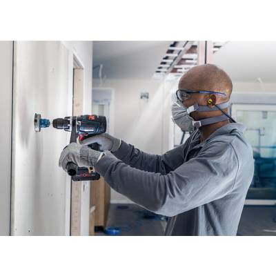 Ein Handwerker bohrt mit einer elektrischen Bohrmaschine ein Loch in eine weiße Wand. Er trägt Schutzbrille, Maske und Handschuhe.