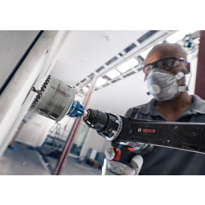 Ein Arbeiter mit Schutzbrille und Maske bohrt mit einer Bohrmaschine ein Loch in eine Wand. Im Hintergrund sind Gerüste sichtbar.