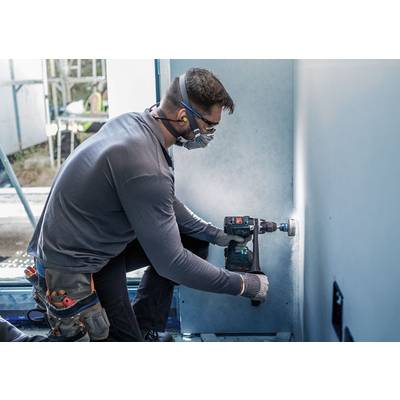 Ein Arbeiter kniet und bohrt ein Loch in eine Wand mit einer Bohrmaschine. Er trägt Schutzbrille, Maske und Gehörschutz.