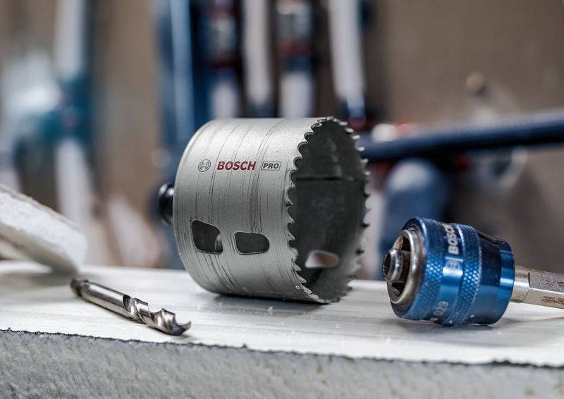 Bohrwerkzeug von Bosch mit Zahnkranz auf Werkbank, daneben ein Bohrer und ein weiteres Werkzeug, fokussiert auf die Metallteile.