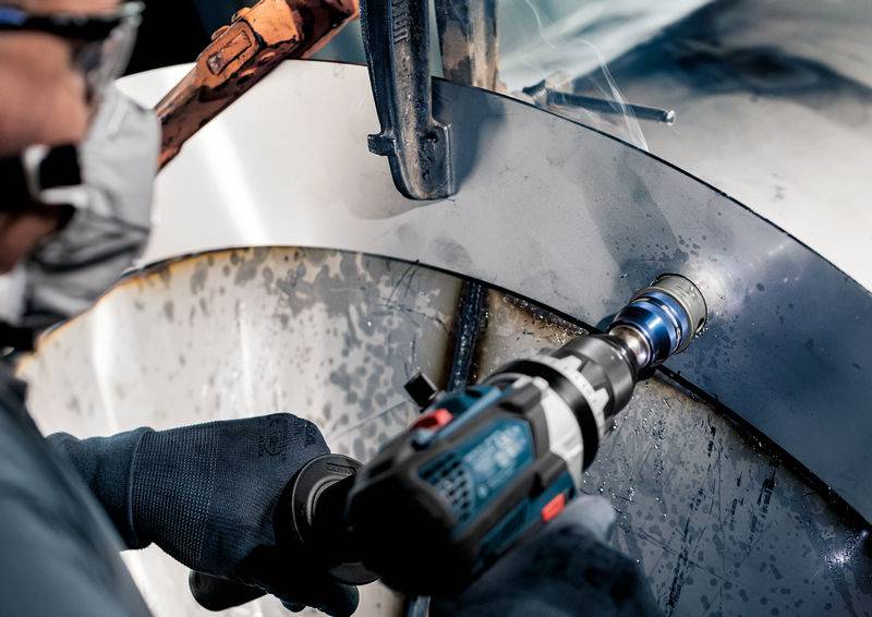 Eine Person arbeitet mit einer Bohrmaschine auf einer Metalloberfläche. Sie trägt Schutzkleidung, einschließlich Handschuhe und Maske.