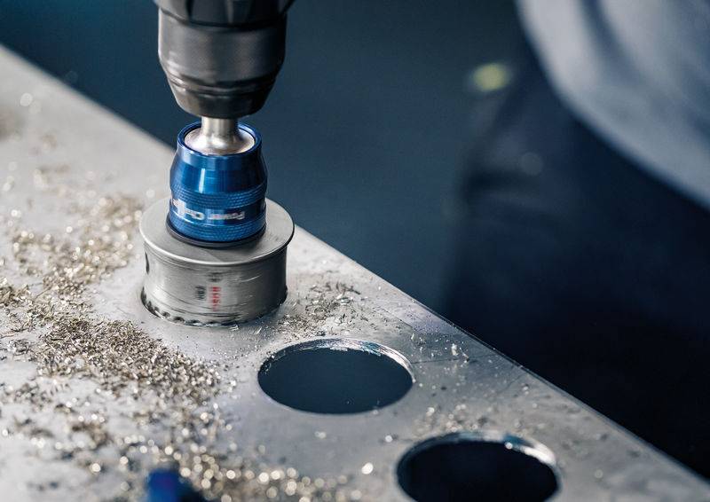 Eine Metallplatte wird mit einem Bohrwerkzeug bearbeitet, das runde Löcher erzeugt. Umgebende Metallspäne sind sichtbar.