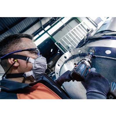 Eine Person mit Schutzbrille und Maske benutzt eine Bohrmaschine an einem großen Metallstück in einer industriellen Umgebung.