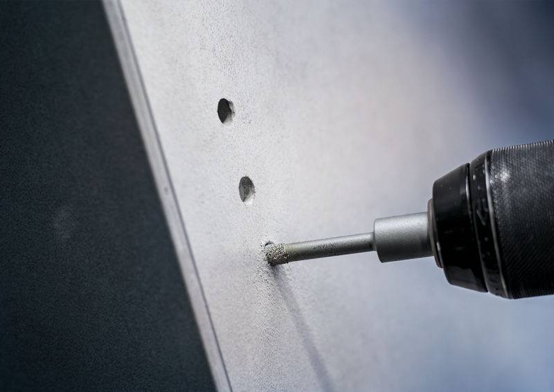 Ein elektrischer Bohrer bohrt in eine weiße Wand mit zwei bereits gebohrten Löchern daneben. Der Bohrer erzeugt Staub.