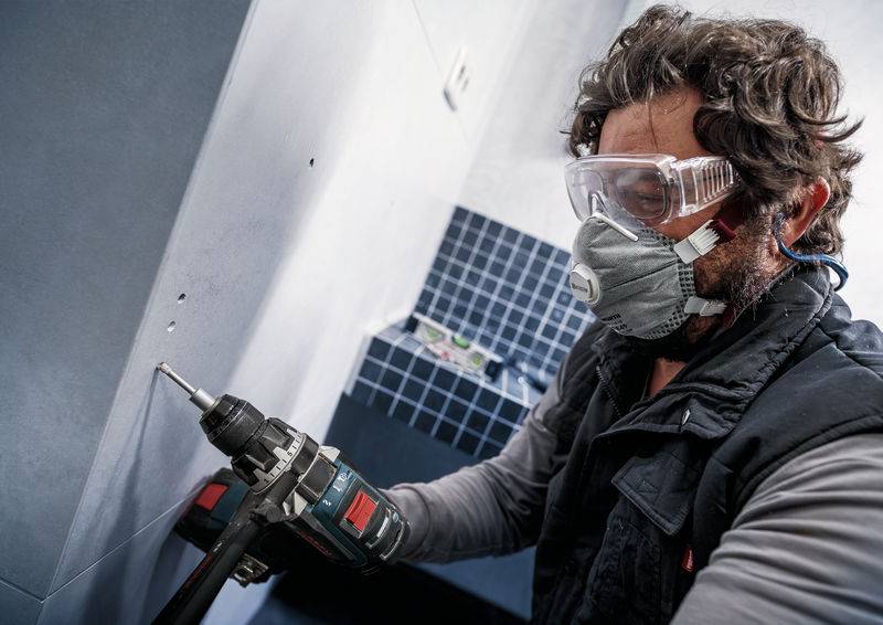 Person bohrt in eine Wand. Sie trägt Schutzbrille und Maske. Hintergrund zeigt blaue Kacheln.