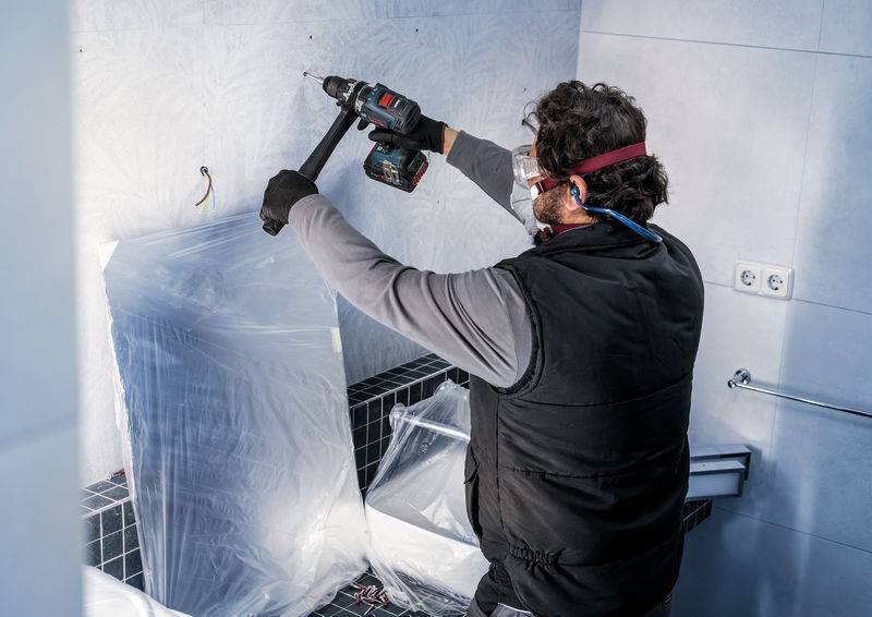 Ein Handwerker montiert eine Halterung in einem Badezimmer. Er trägt Schutzbrille und Handschuhe, nutzt eine Bohrmaschine.