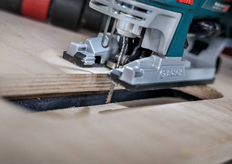 Eine Stichsäge schneidet in ein Holzbrett. Die Säge ist grün und grau, präzises Arbeiten an einer rechteckigen Aussparung.