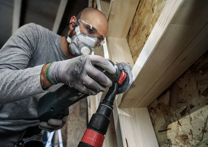 Ein Handwerker mit Schutzbrille und Atemmaske schleift ein Holzbrett mit einem Elektrowerkzeug in einer Werkstattumgebung.