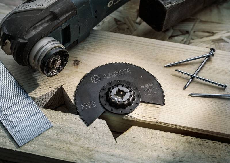 Multifunktionswerkzeug neben Sägeblatt und Nägeln auf einem Holzbalken. Das Sägeblatt trägt die Aufschrift 'Bosch PRO'.