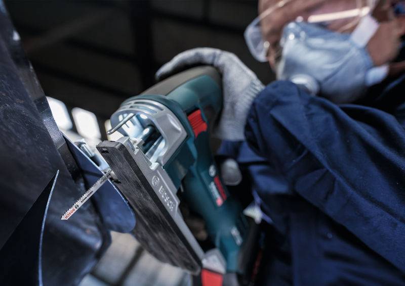 Person mit Schutzbrille und Handschuhen verwendet eine elektrische Säge, um Metall zu schneiden. Fokus auf die Sägeklinge.