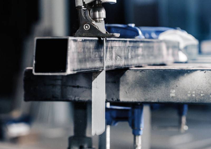 Eine Bandsäge schneidet ein quadratisches Metallrohr auf einer Werkbank. Die Umgebung scheint industriell oder eine Werkstatt zu sein.