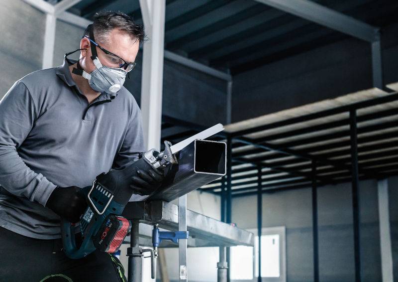 Mann in Schutzkleidung schneidet mit einer Elektrosäge ein Metallrohr in einer Werkstatt.