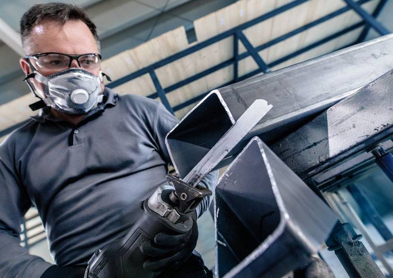 Ein Arbeiter mit Schutzbrille und Maske verwendet ein Elektrowerkzeug, um Metall zu schneiden. Im Hintergrund eine Stahlkonstruktion.