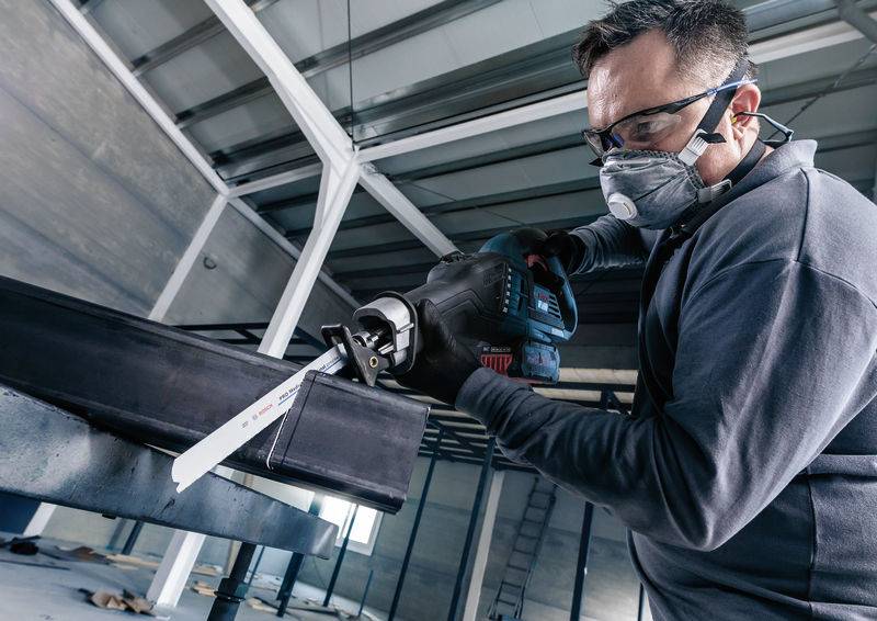 Ein Mann trägt Schutzbrille und Maske, während er mit einer elektrischen Säge an einem Metallbalken in einer Werkstatt arbeitet.