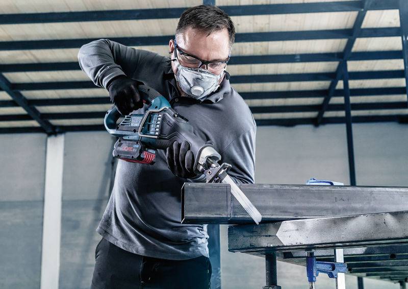 Ein Arbeiter mit Schutzbrille und Maske schneidet ein Metallrohr mit einer Elektrosäge in einer Werkstattumgebung.