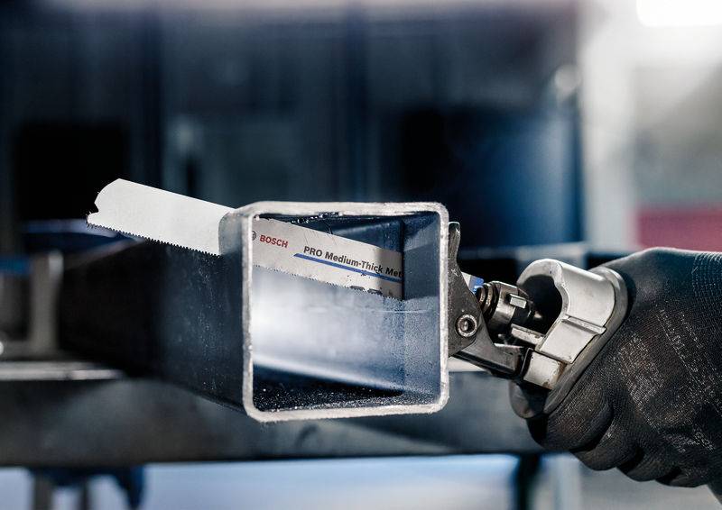 Ein schwarzhandschuhter Arbeiter sägt mit einer Metallsäge einen rechteckigen Metallbalken durch.