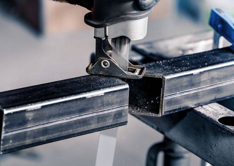 Eine Nahaufnahme einer Person, die mit einer Säge Metallrohre schneidet. Metallspäne sind sichtbar. Die Umgebung wirkt industriell.