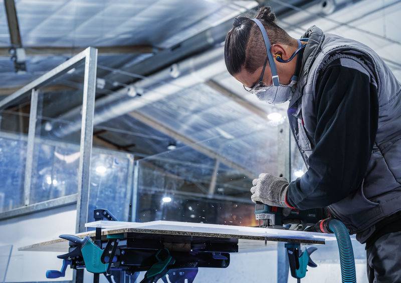 Eine Person schleift in einer Werkstatt mit Schutzbrille, Maske und Gehörschutz ein Holzstück auf einer Werkbank.