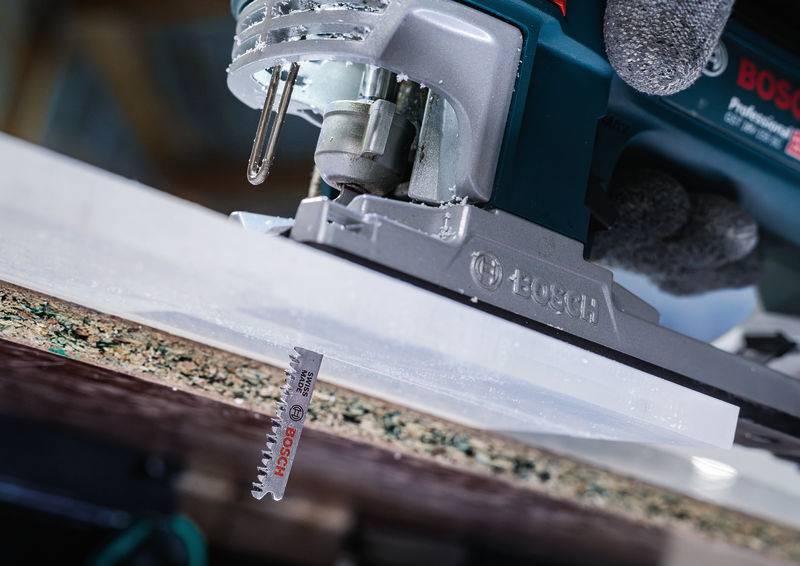 Eine elektrische Stichsäge schneidet durch eine weiße Kunststoffplatte. Die Säge hat einen blaugrauen Griff und ein Sägeblatt mit Zähnen.