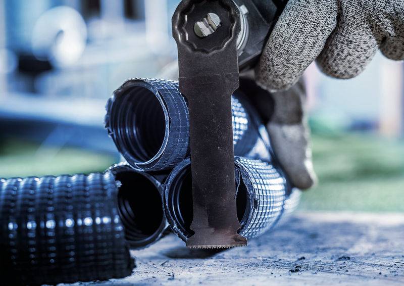 Handschuhhand schneidet schwarzes Metallrohrbündel auf Baustelle. Nahaufnahme der Arbeit mit spezialisierter Schneidklinge.
