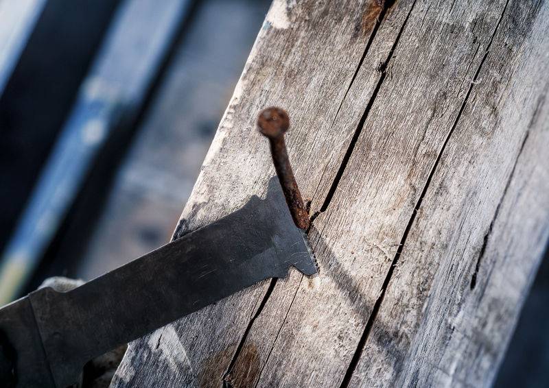 Nahaufnahme eines alten Holzbalkens mit einem hervorstehenden rostigen Nagel, neben dem eine Säge liegt.