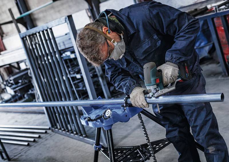 Eine Person trägt Schutzkleidung und schleift mit einem Elektrowerkzeug ein Metallrohr in einer Werkstatt.
