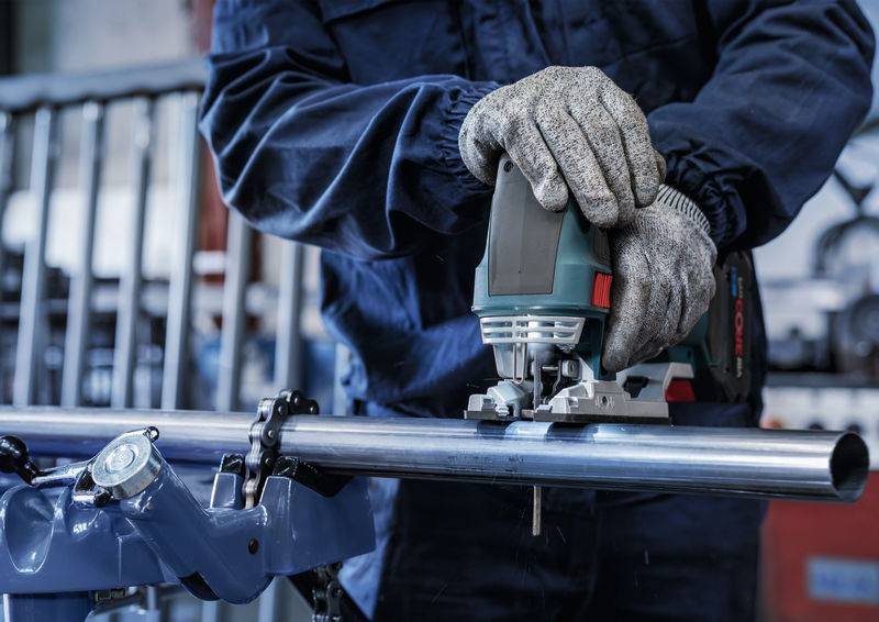 Eine Person in Arbeitskleidung schneidet mit einer Stichsäge ein Metallrohr. Ihre Hände tragen Schutzhandschuhe.
