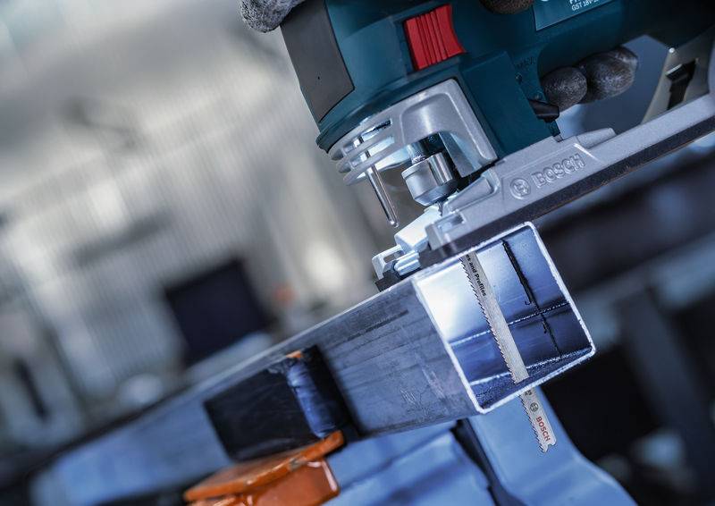 Eine elektrische Säge schneidet ein Metallprofil, das in einem Schraubstock eingespannt ist. Die Szene spielt in einer Werkstatt.